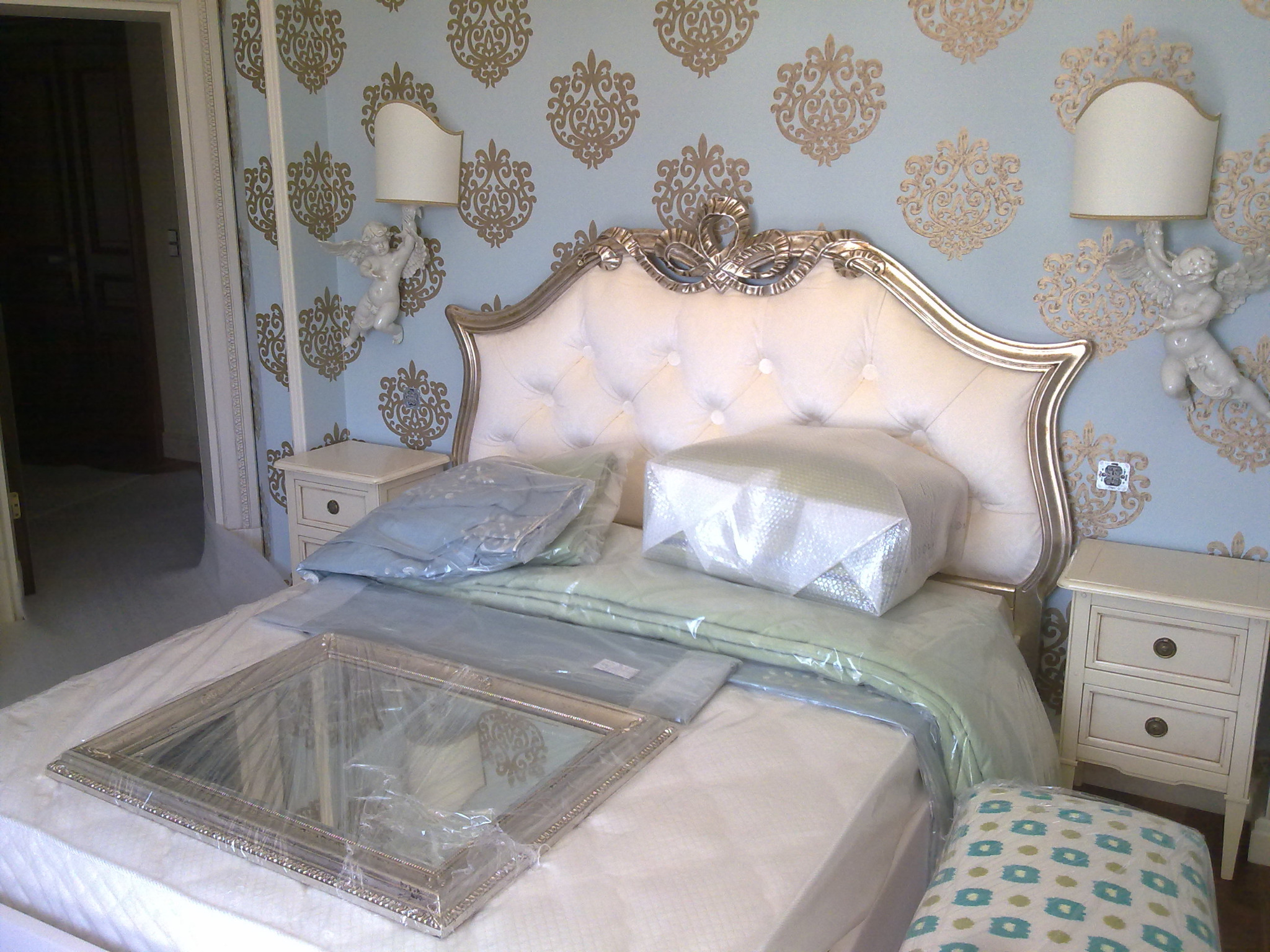 Pokój w stylu glamour przed wykończeniem: łóżko z tapicerowanym zagłówkiem w kolorze kremowym, szafki nocne, dekoracyjne kinkiety z aniołkami, ściany z ornamentami w odcieniach złota i błękitu...