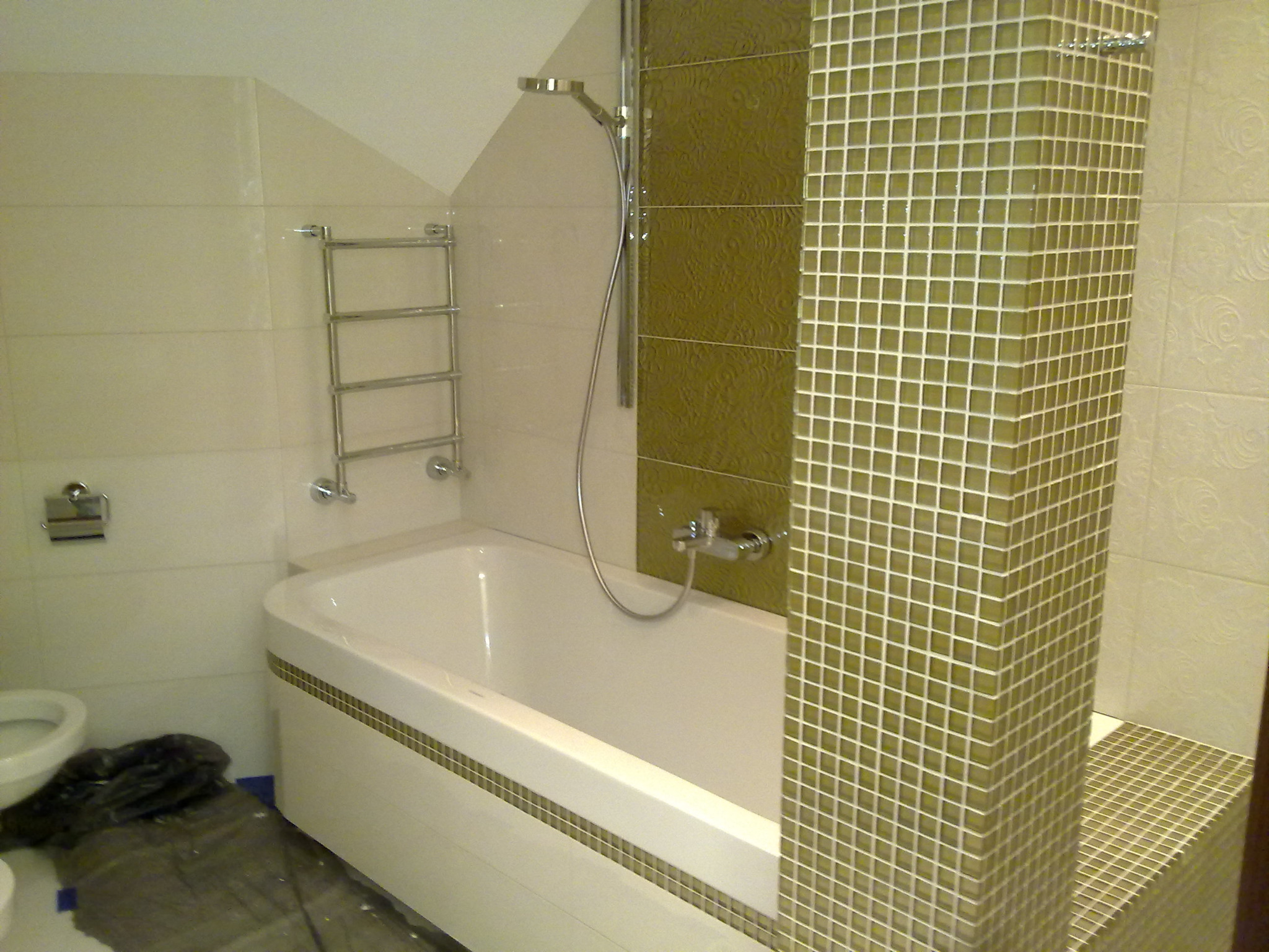 Wykończona łazienka z białą wanną obudowaną mozaiką, chromowana bateria i deszczownica, ściany wyłożone płytkami w odcieniach beżu i oliwki, widoczne zabezpieczenia podłogi folią malarską.