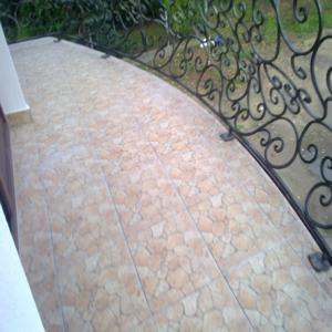 Wyłożony jasnymi płytkami balkon z ozdobną, kutą balustradą. Płytki ułożone wzdłuż łuku balkonu, widoczne spoiny. W tle zieleń ogrodu.