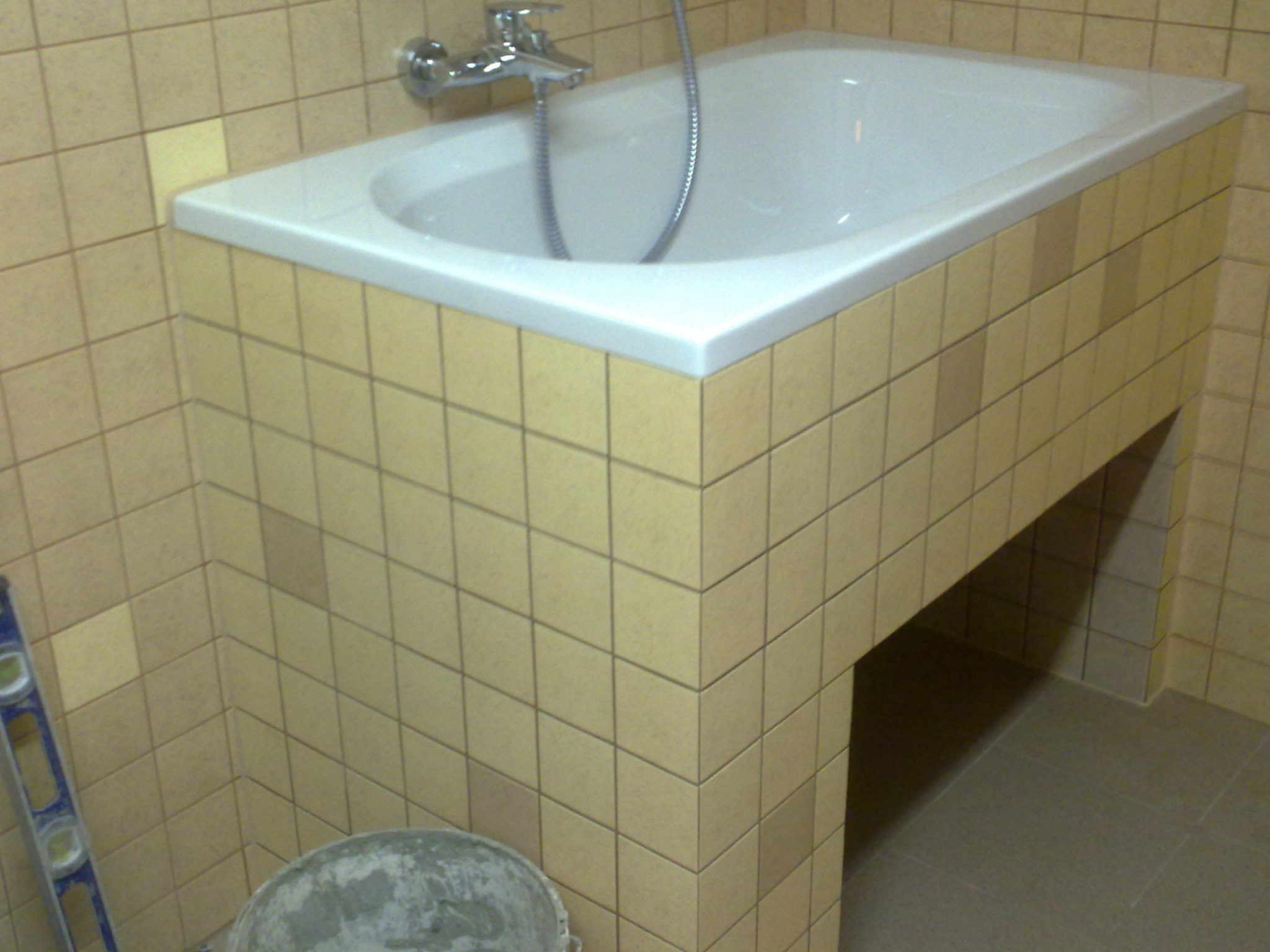 Obudowana beżowymi płytkami wanna z baterią, widoczny poziomica i wiadro z zaprawą, fragment wykończonej łazienki.