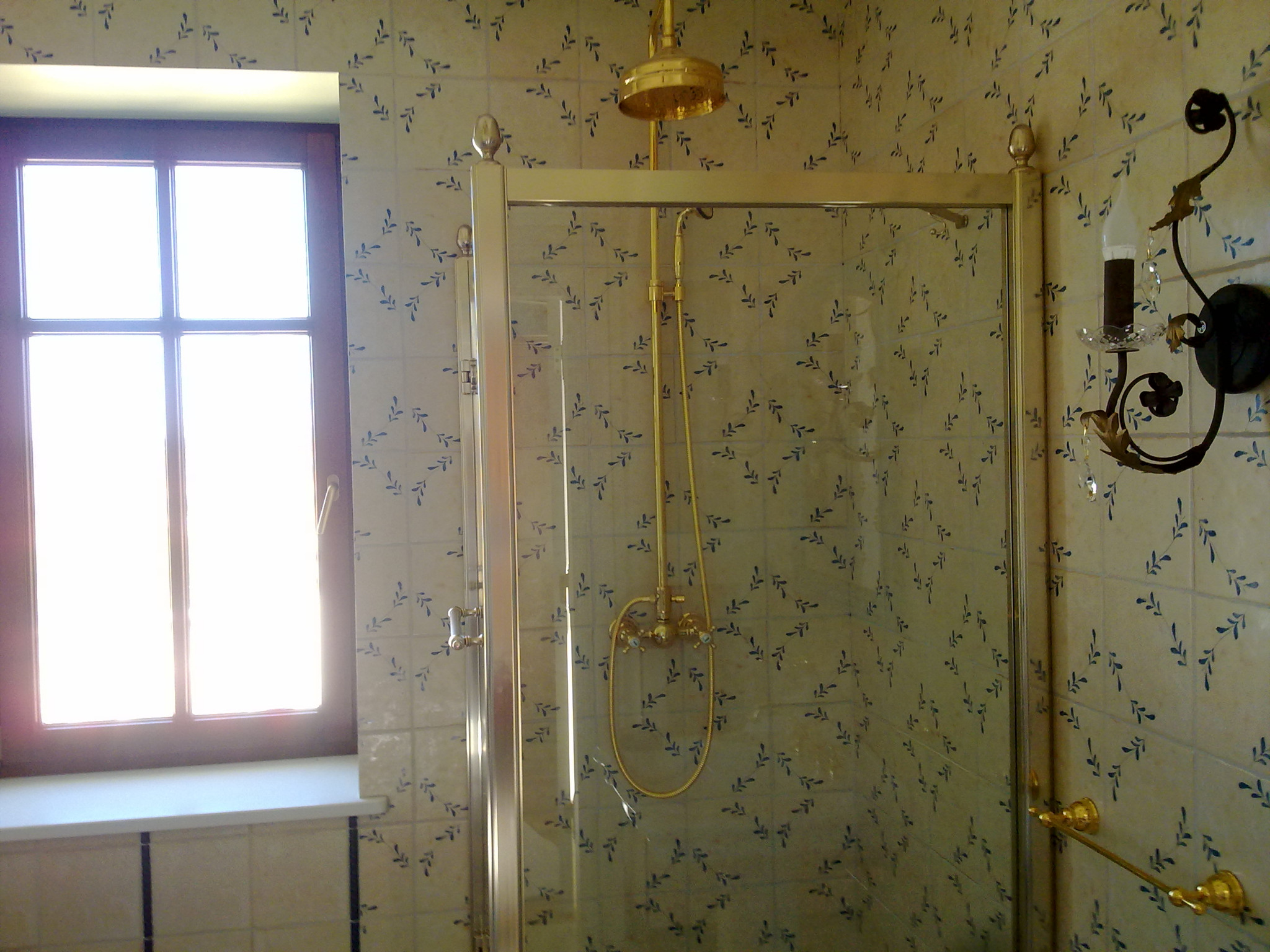 Wykończona łazienka z kabiną prysznicową w złotej ramie, baterią prysznicową w stylu retro, kremowymi płytkami z motywem niebieskich listków, oświetlona światłem z okna.