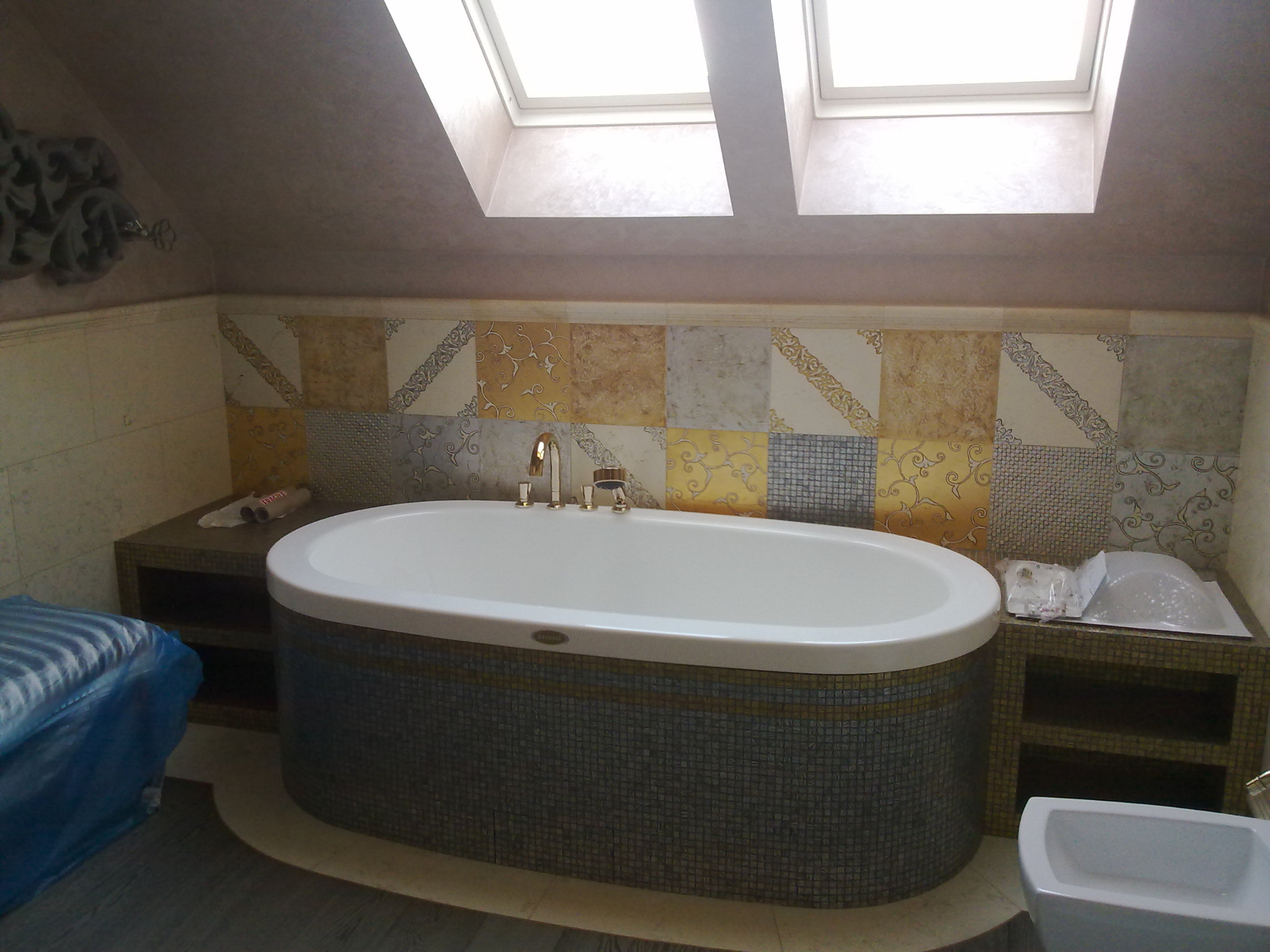 Wykończona łazienka na poddaszu z owalną wanną obudowaną mozaiką, wzorzyste płytki na ścianie, dwa okna dachowe i dekoracyjny ornament na ścianie.