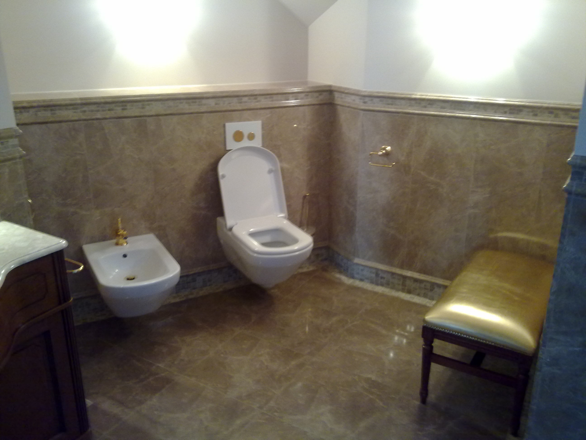 Elegancka łazienka w odcieniach beżu z marmurowymi płytkami, wiszącą toaletą, bidetem z pozłacaną armaturą i ławką z błyszczącym, złotym siedziskiem.