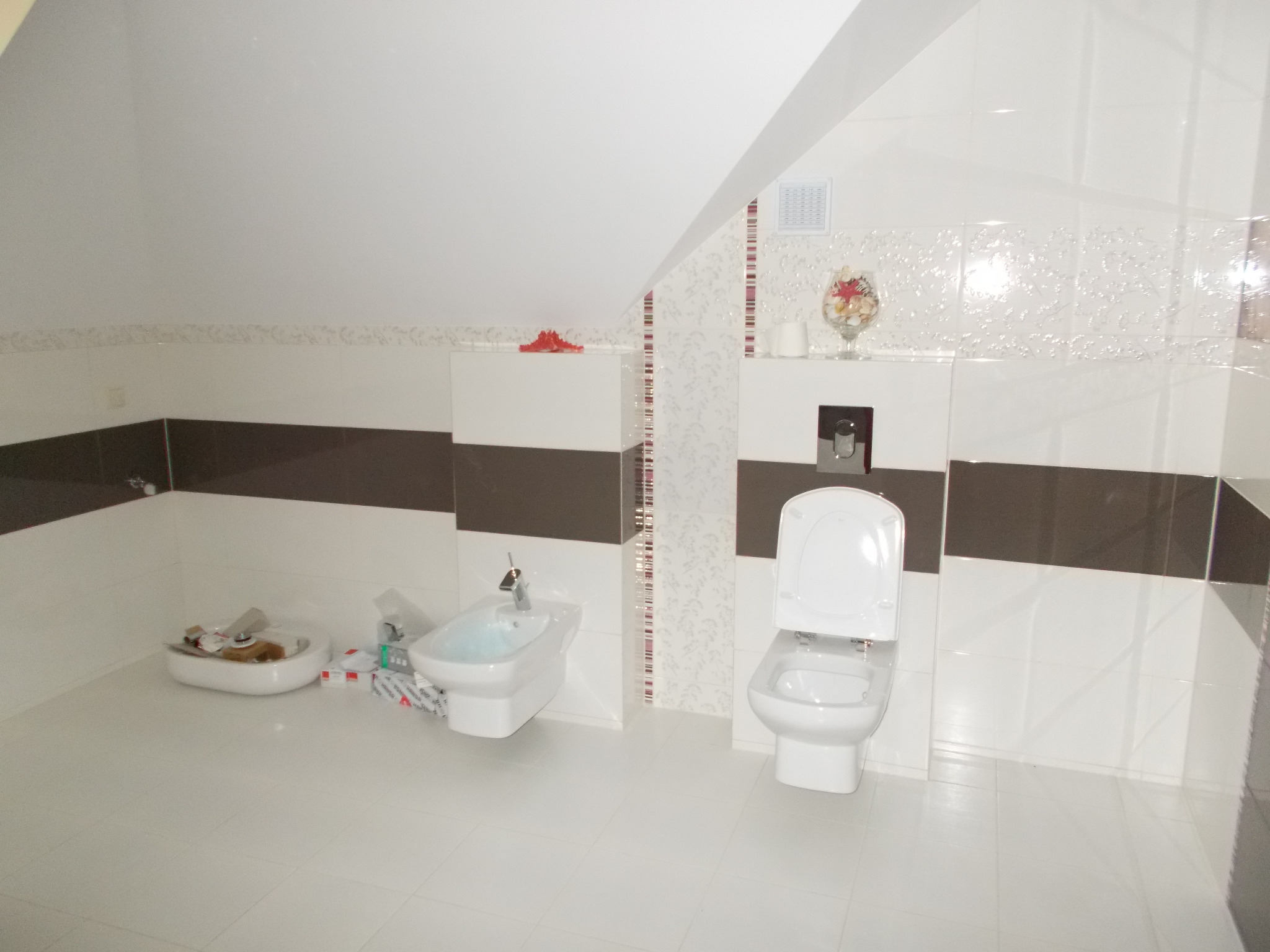 Biała łazienka na poddaszu z umywalką, bidetem i toaletą, wykończona płytkami w odcieniach bieli i brązu, z dekoracyjnym wzorem na ścianie i akcesoriami łazienkowymi.