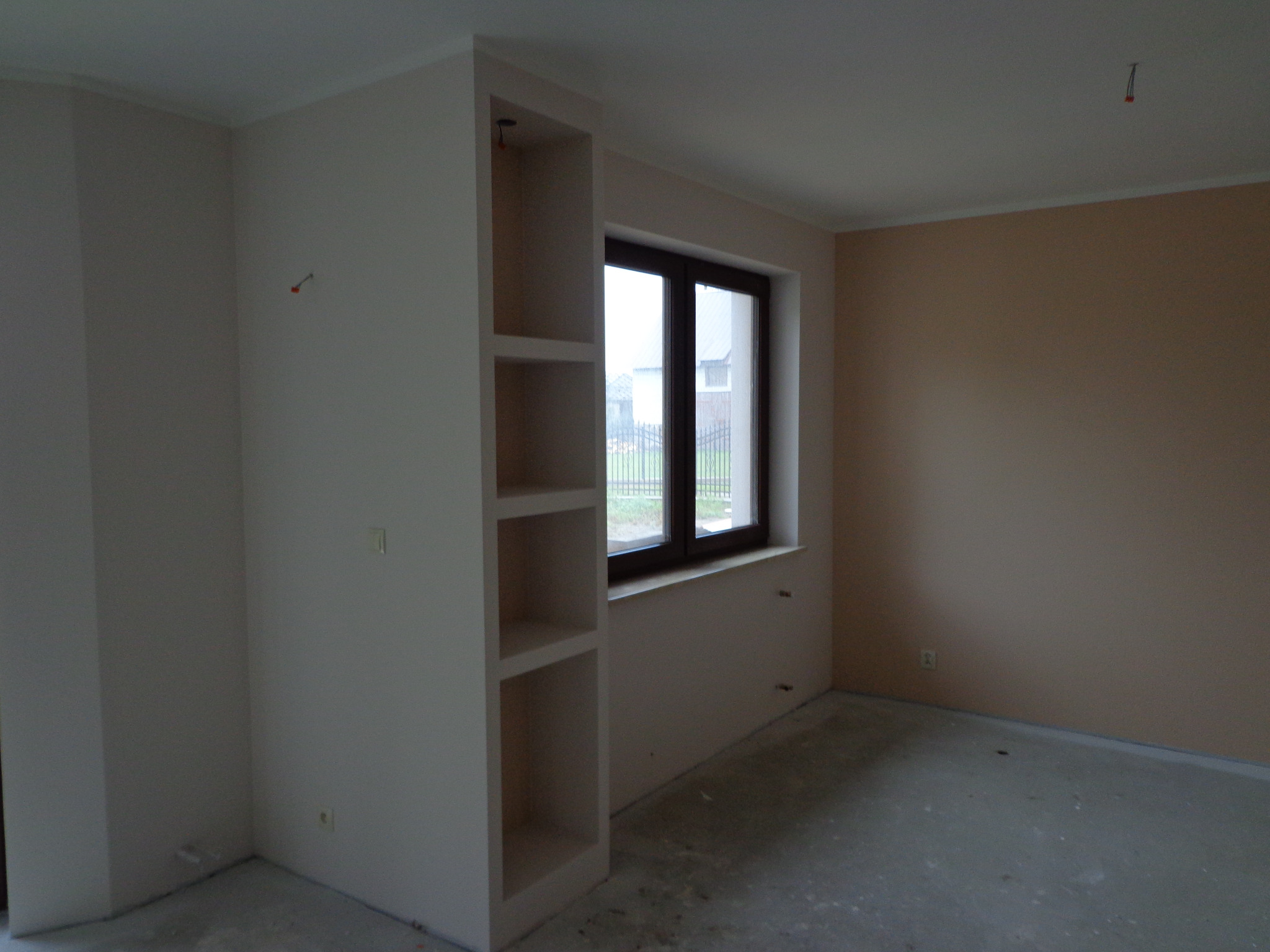 Pokój w trakcie wykańczania, z oknem i wnęką z półkami z płyty kartonowo-gipsowej, ściany częściowo pomalowane na beżowo, surowa podłoga, widoczne wystające kable elektryczne.