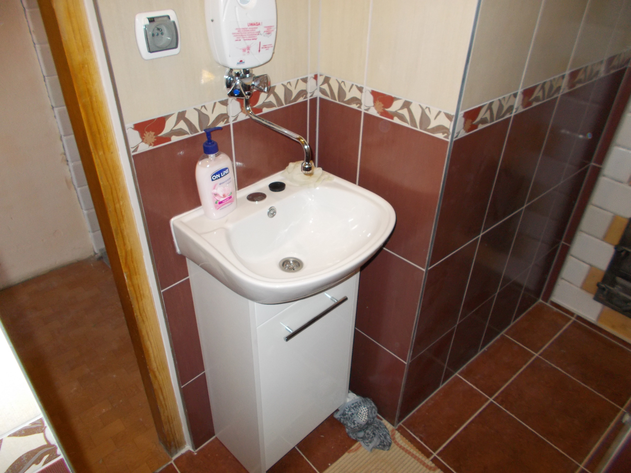 Mała umywalka z szafką w łazience z brązowymi płytkami, dozownik mydła i grzejnik przepływowy nad umywalką.