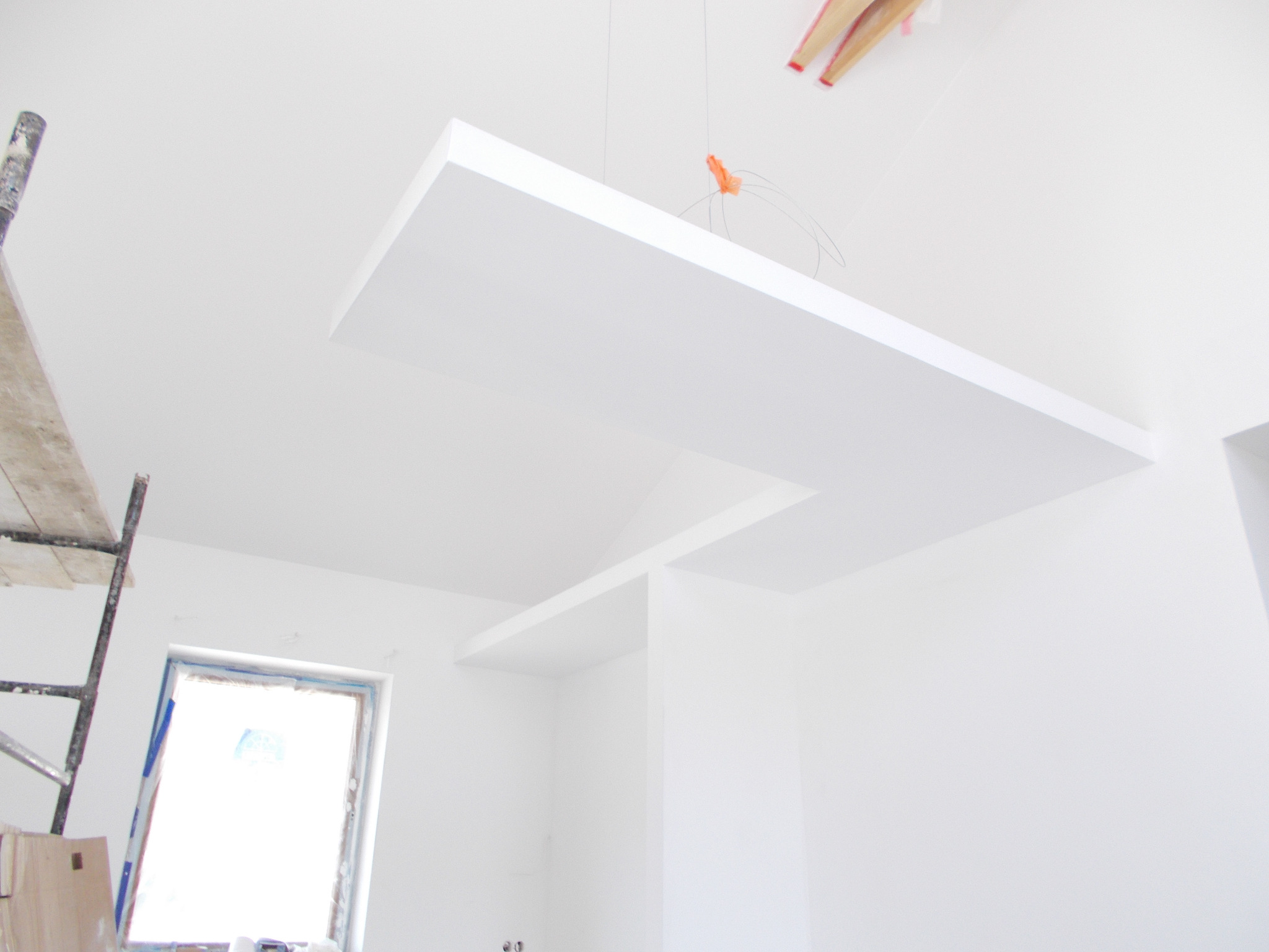 Białe, minimalistyczne wnętrze w trakcie wykończenia z podwieszanym sufitem z płyt kartonowo-gipsowych, widoczny fragment rusztowania malarskiego przy oknie.