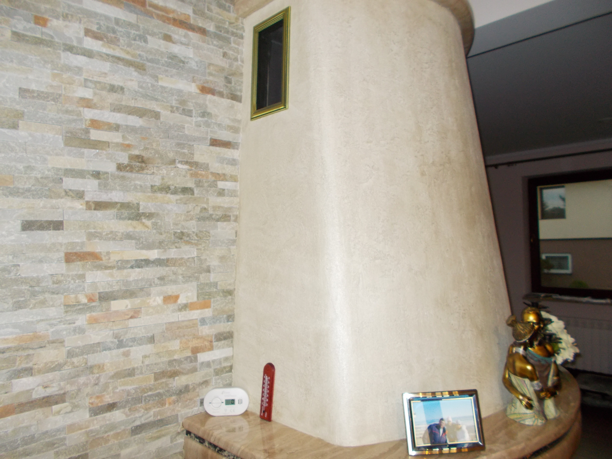 Obudowa kominka z beżową, teksturowaną powierzchnią, dekorowana ramką z czarną kratką, przylegająca do ściany wyłożonej kamienną mozaiką w odcieniach szarości i beżu.