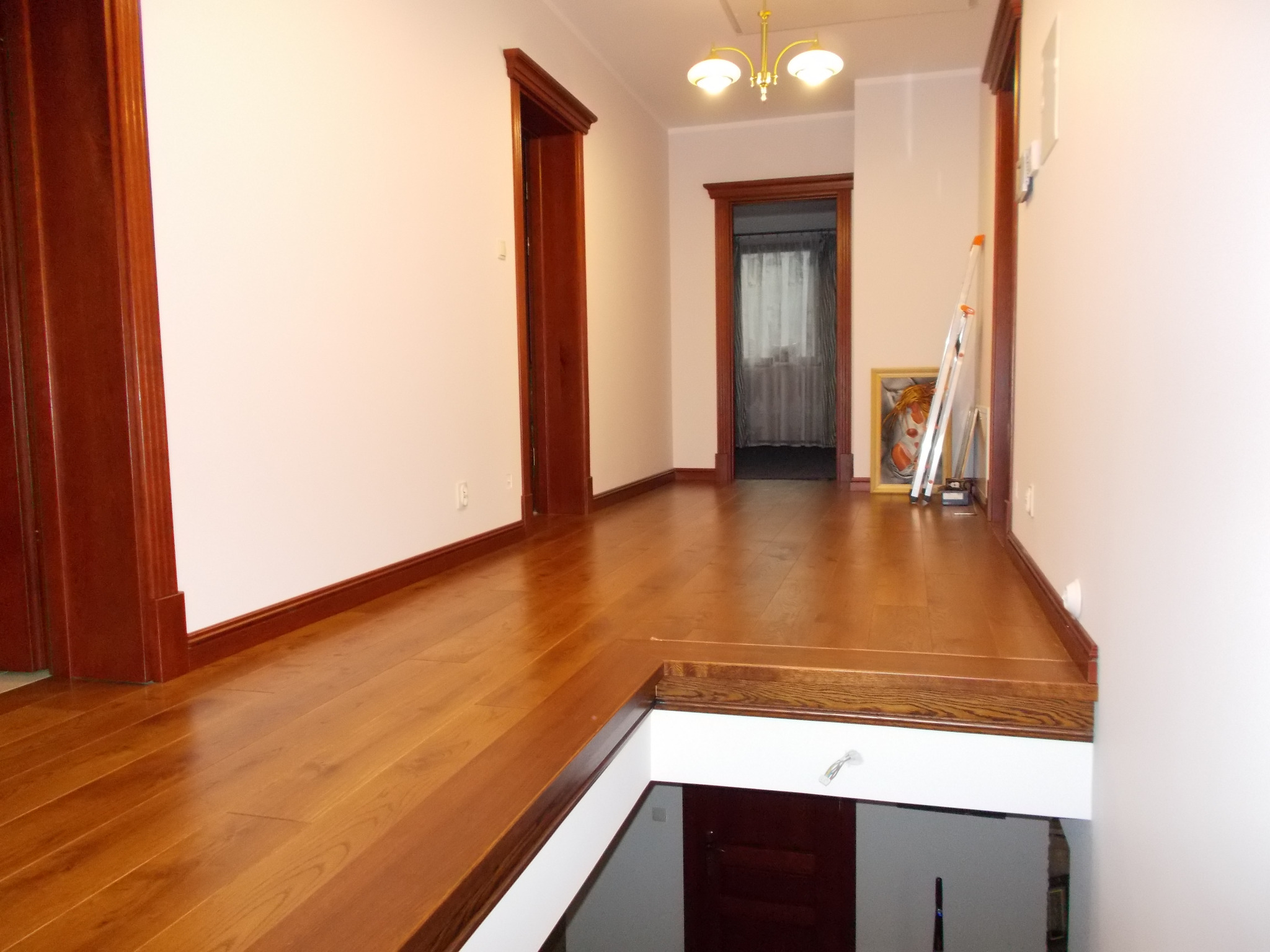 Jasne wnętrze domu z drewnianą podłogą, widoczne drzwi z ciemnego drewna, schody prowadzące w dół i drabina oparta o ścianę w tle.