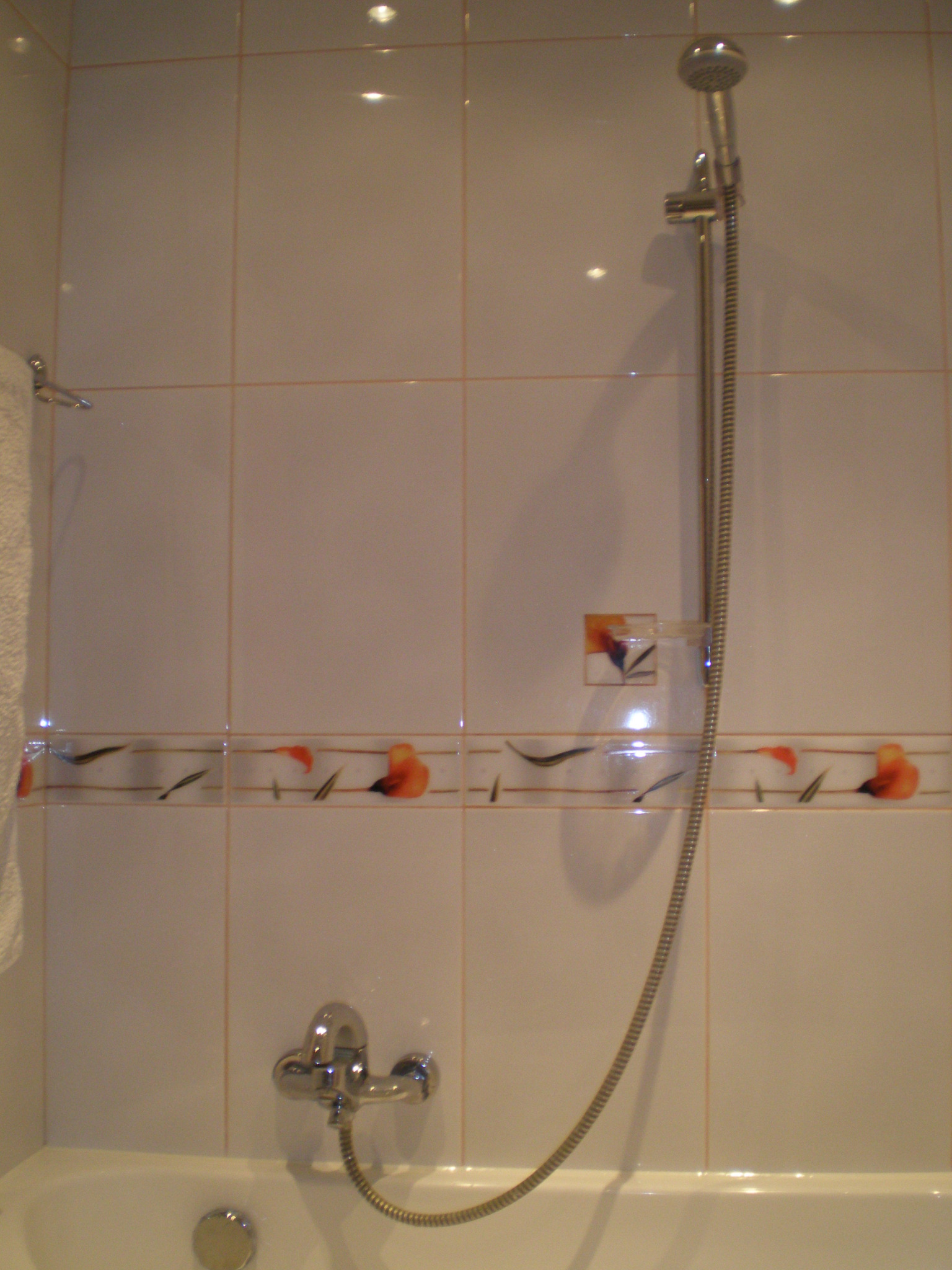 Białe płytki ścienne z dekoracyjnym motywem kwiatowym w łazience, bateria wannowa z chromowaną słuchawką prysznicową na regulowanym drążku.