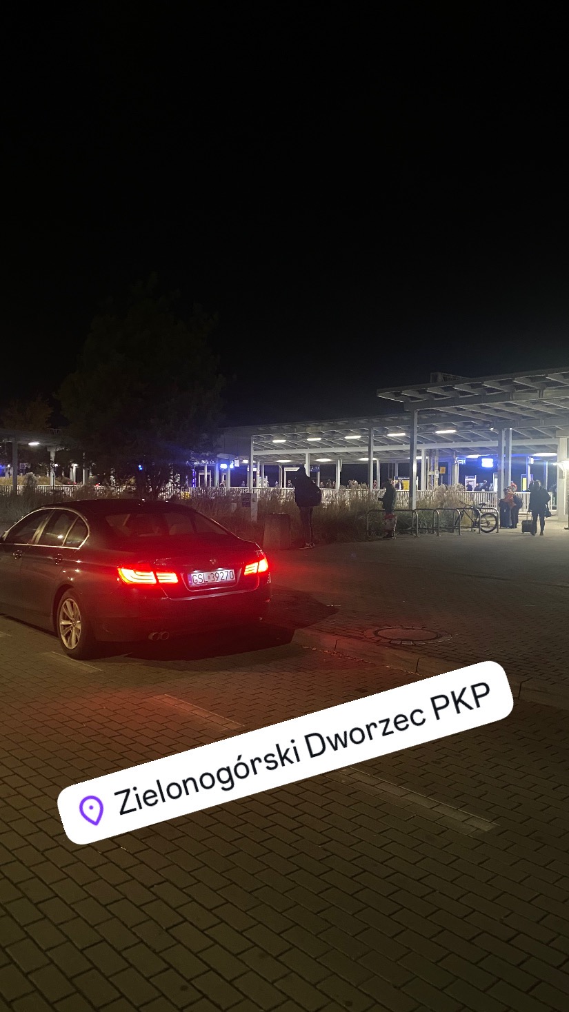 Ciemnobrązowy samochód osobowy z włączonymi światłami stopu stoi na parkingu przed dworcem PKP w Słupsku nocą.