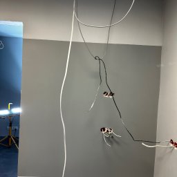 RBK Construction - Ściana pomalowana na dwa odcienie szarości z wystającymi przewodami elektrycznymi i puszkami, widoczny fragment sufitu podwieszanego i lampa robocza w tle.