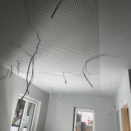 RBK Construction - Montaż perforowanego sufitu podwieszanego w szarym pomieszczeniu z wystającymi kablami elektrycznymi, widoczne dwa okna i przygotowane materiały na parapecie.
