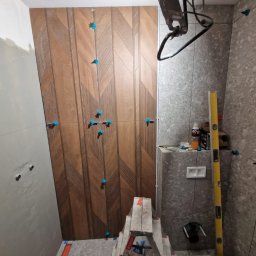 RBK Construction - Montaż płytek drewnopodobnych w łazience, widoczne niebieskie krzyżyki dystansowe, poziomica i drabina.