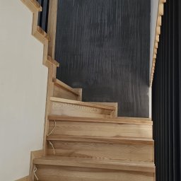 RBK Construction - Jasne drewniane schody z czarną metalową balustradą na tle ściany z szarą dekoracyjną fakturą.