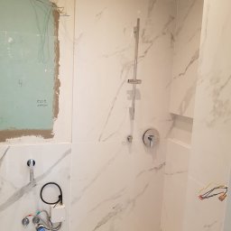 RBK Construction - Łazienka w trakcie wykańczania: białe płytki imitujące marmur, zamontowana bateria prysznicowa, widoczne instalacje wodne i elektryczne przed zabudową, fragment brodzika, ściana przygotowana...
