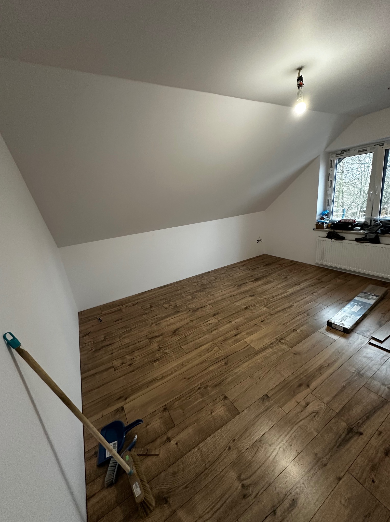 Podłoga z paneli drewnopodobnych w pokoju na poddaszu, częściowo ułożona, widoczne narzędzia do sprzątania i paczka paneli przy oknie.