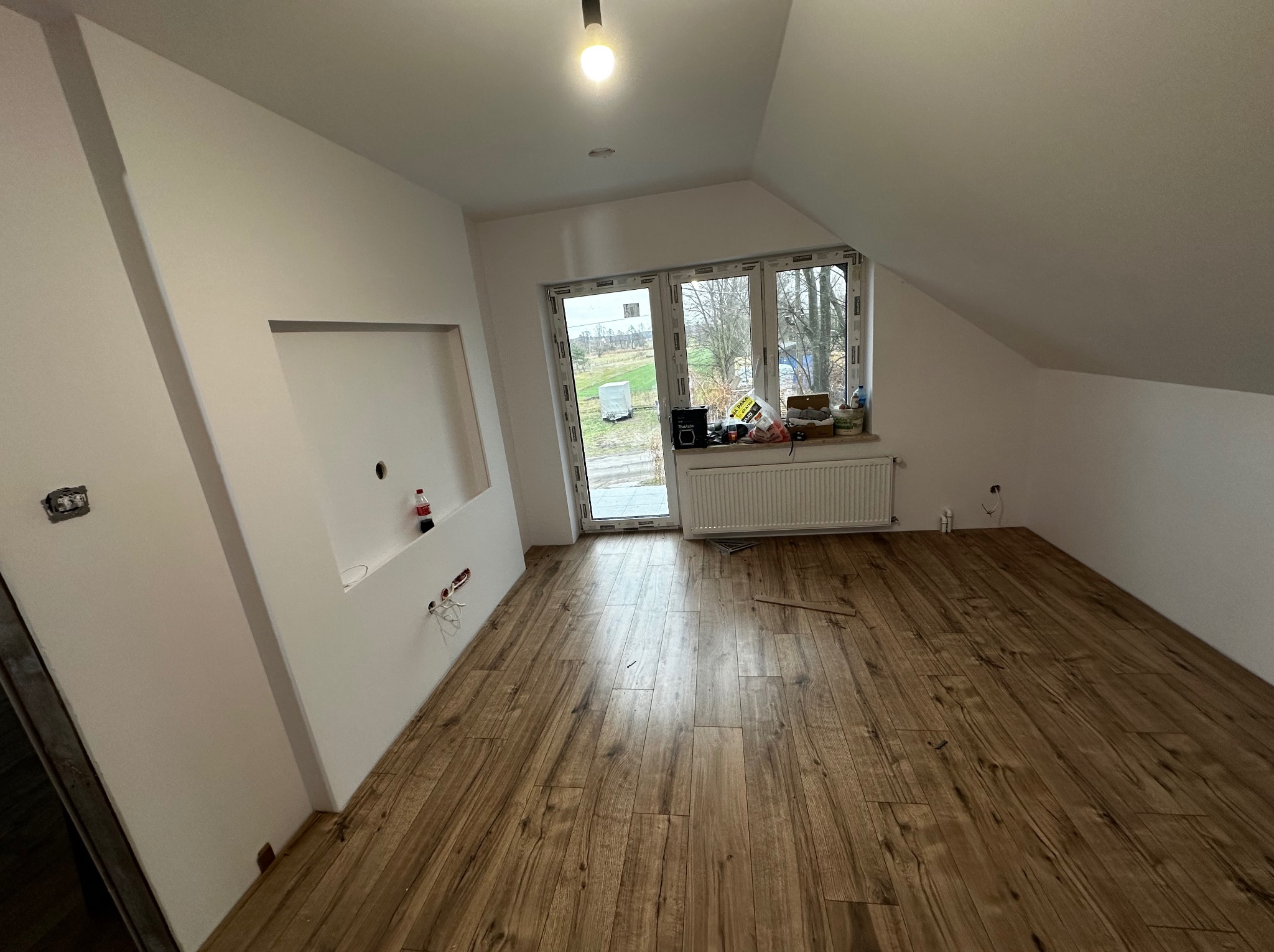 Poddasze w trakcie prac wykończeniowych: widok na ułożone panele podłogowe imitujące drewno, białe ściany z przygotowanymi gniazdkami elektrycznymi, okno dachowe z widokiem na zewnątrz i grzejnik...