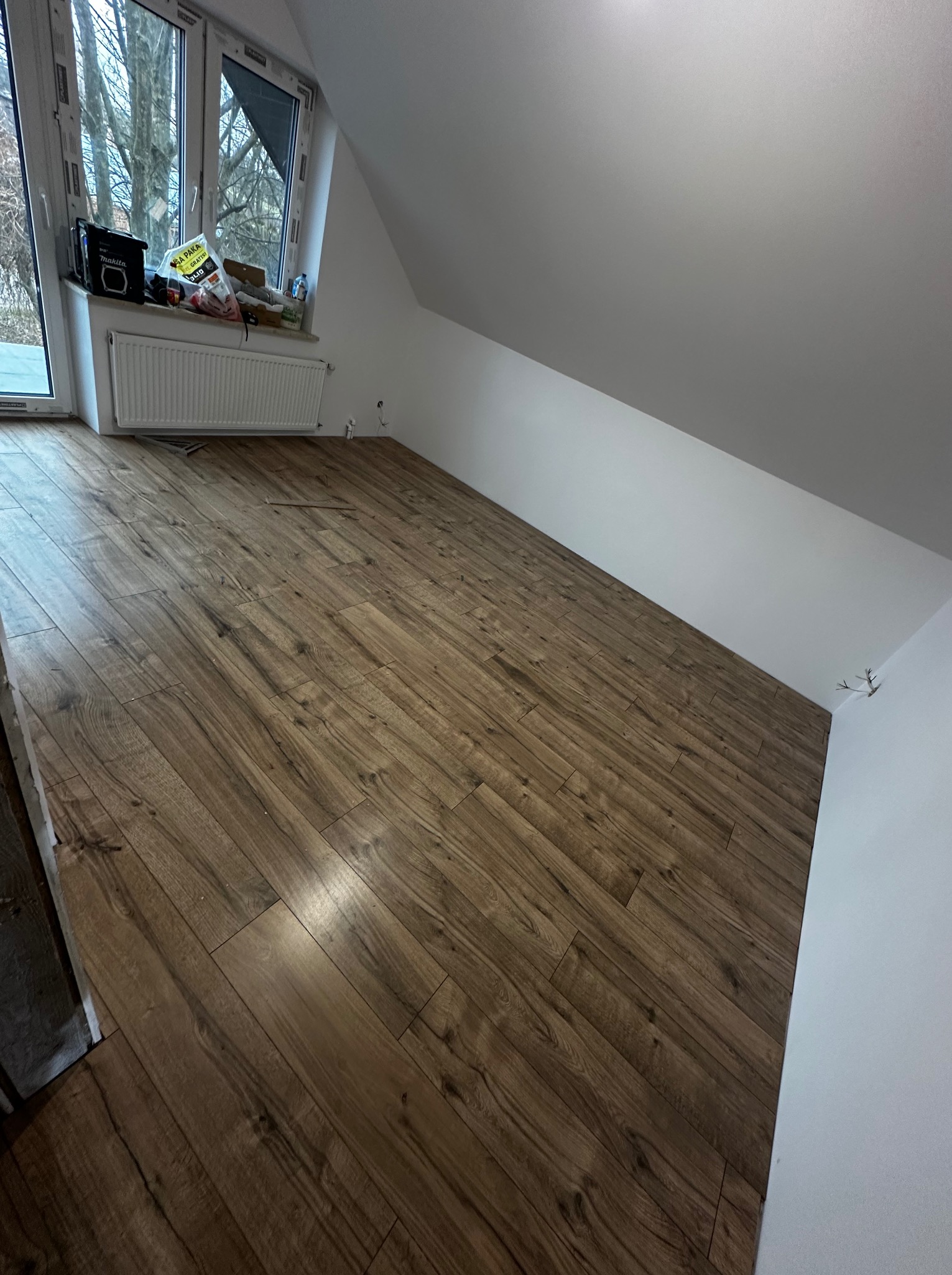 Ukończone układanie paneli podłogowych w pokoju na poddaszu, widoczne okno dachowe i grzejnik.