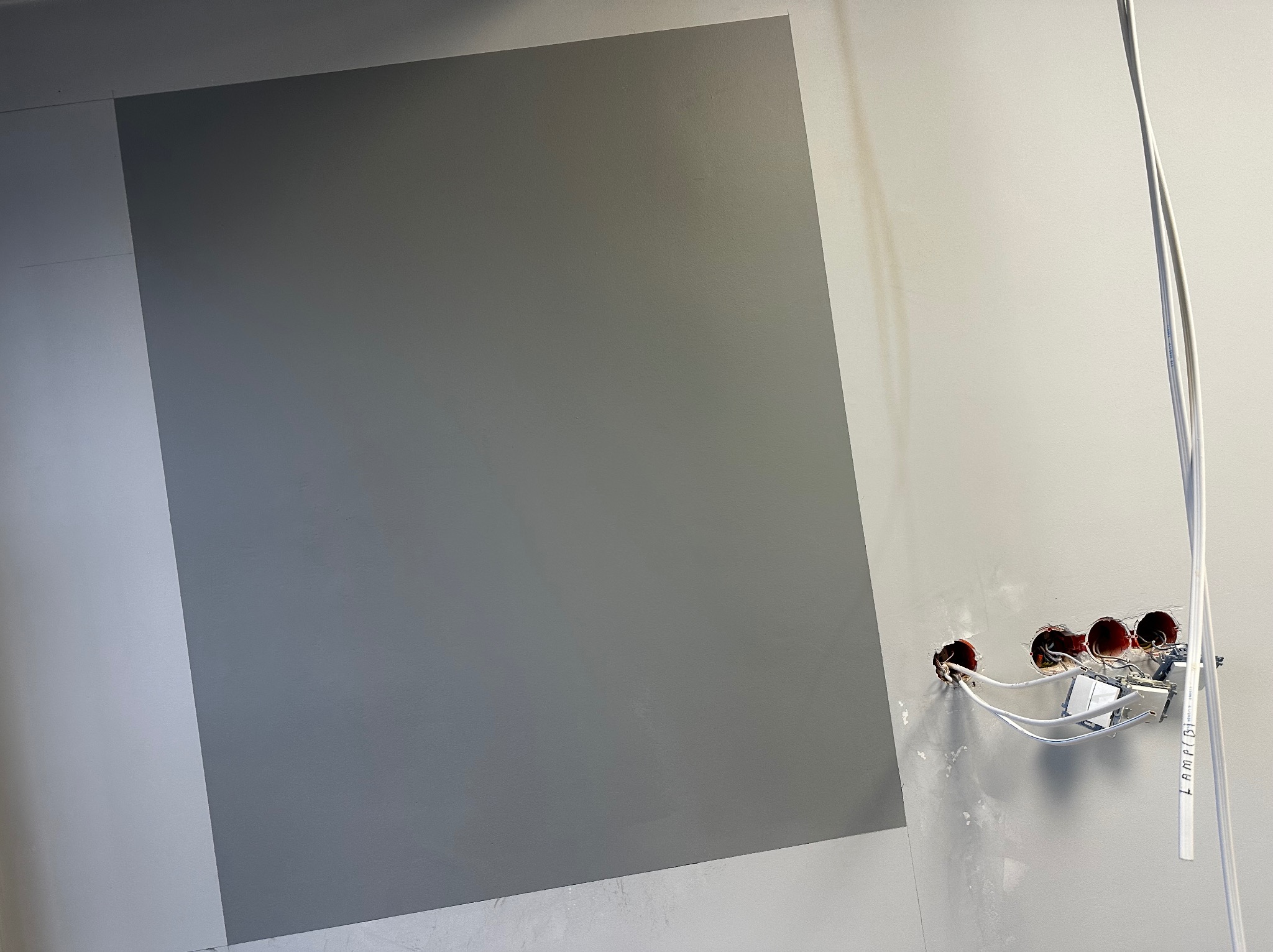 Fragment ściany pomalowanej na dwa odcienie szarości, z widoczną instalacją elektryczną w trakcie montażu gniazdek.