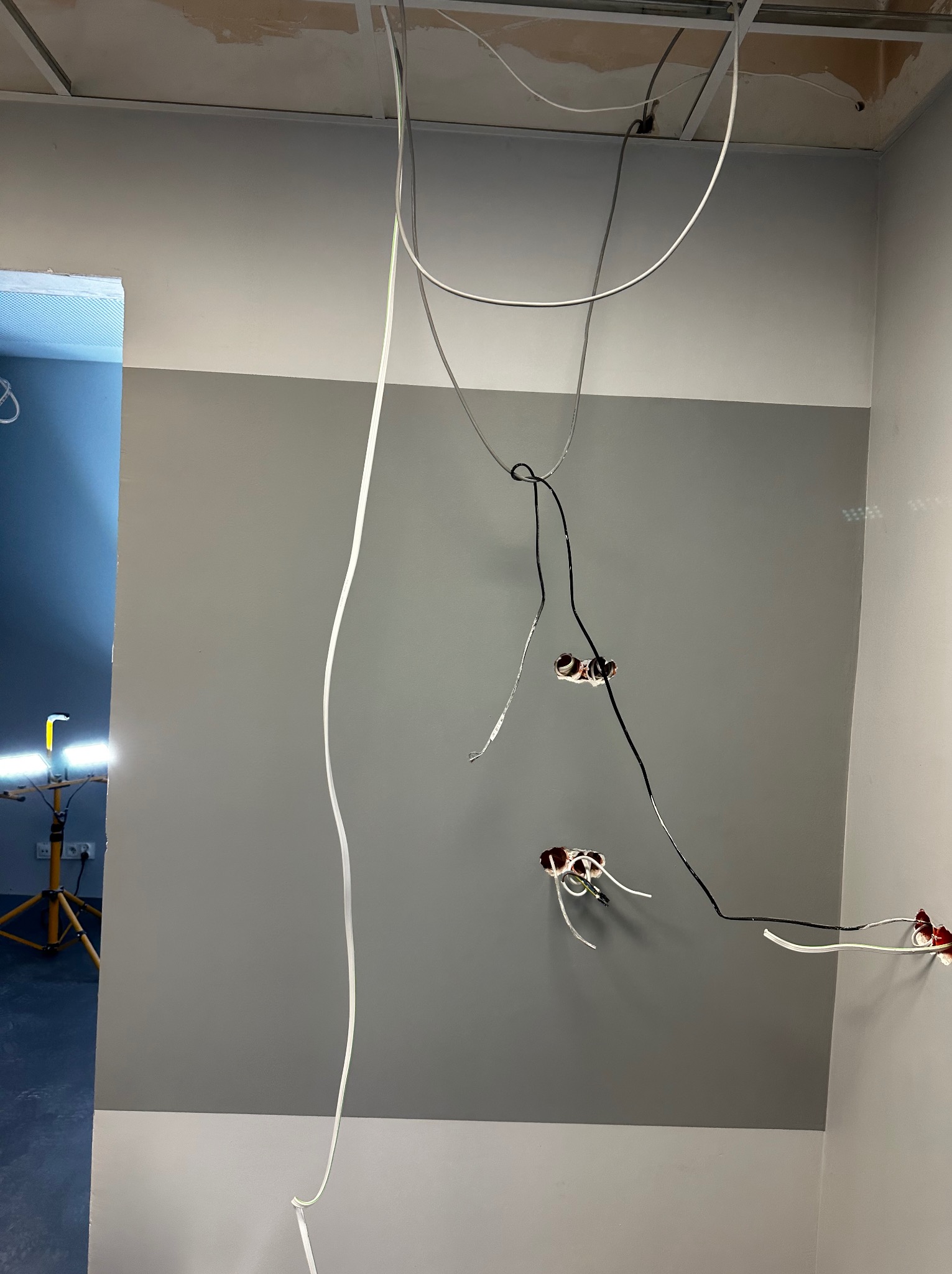 Ściana pomalowana na dwa odcienie szarości z wystającymi przewodami elektrycznymi i puszkami, widoczny fragment sufitu podwieszanego i lampa robocza w tle.