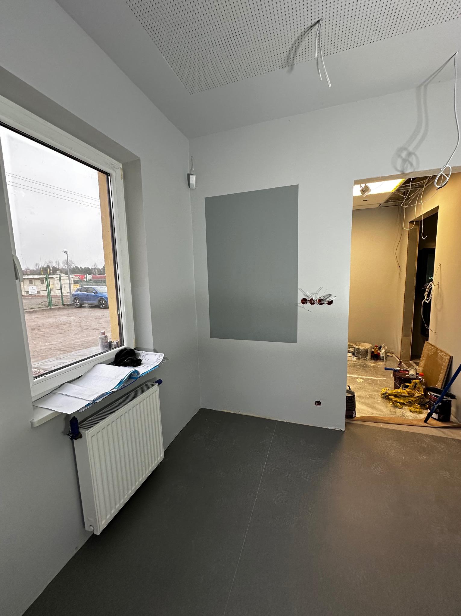 Wnętrze w trakcie odświeżania: szare ściany, gotowe gładzie, widoczne przewody elektryczne, okno z widokiem na zewnątrz, grzejnik pod oknem z planami i czapką, ciemna podłoga, bałagan w sąsiednim...