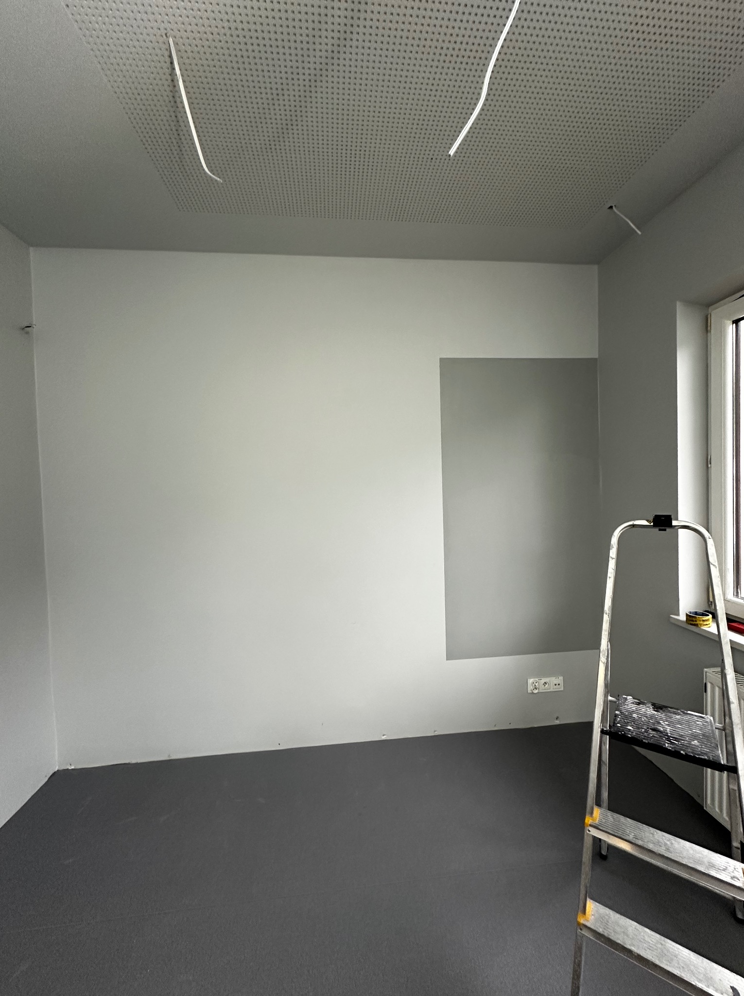 Przygotowane do malowania wnętrze pokoju z szarą podłogą, białą ścianą z fragmentem pomalowanym na szaro i aluminiową drabiną przystawioną do ściany przy oknie.