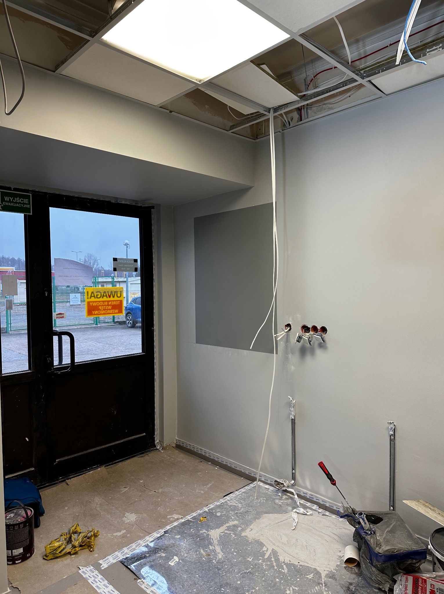 Wnętrze w trakcie malowania, widoczne puszki z farbą, narzędzia malarskie, odsłonięty sufit podwieszany i wyprowadzone instalacje elektryczne na ścianie.