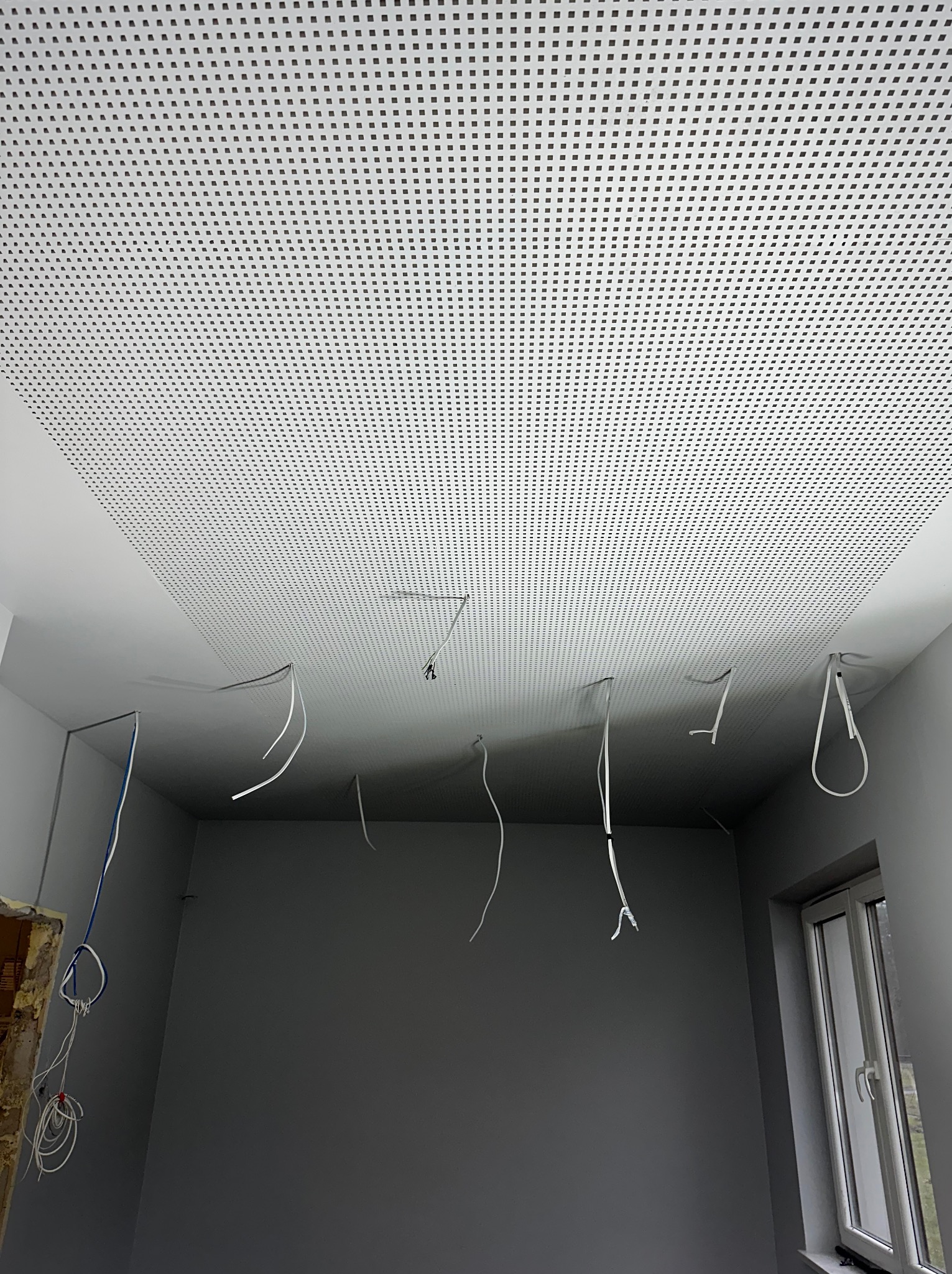 Montaż sufitu podwieszanego z perforowanymi panelami w szarym pomieszczeniu, widoczne przewody instalacji elektrycznej i fragment okna.