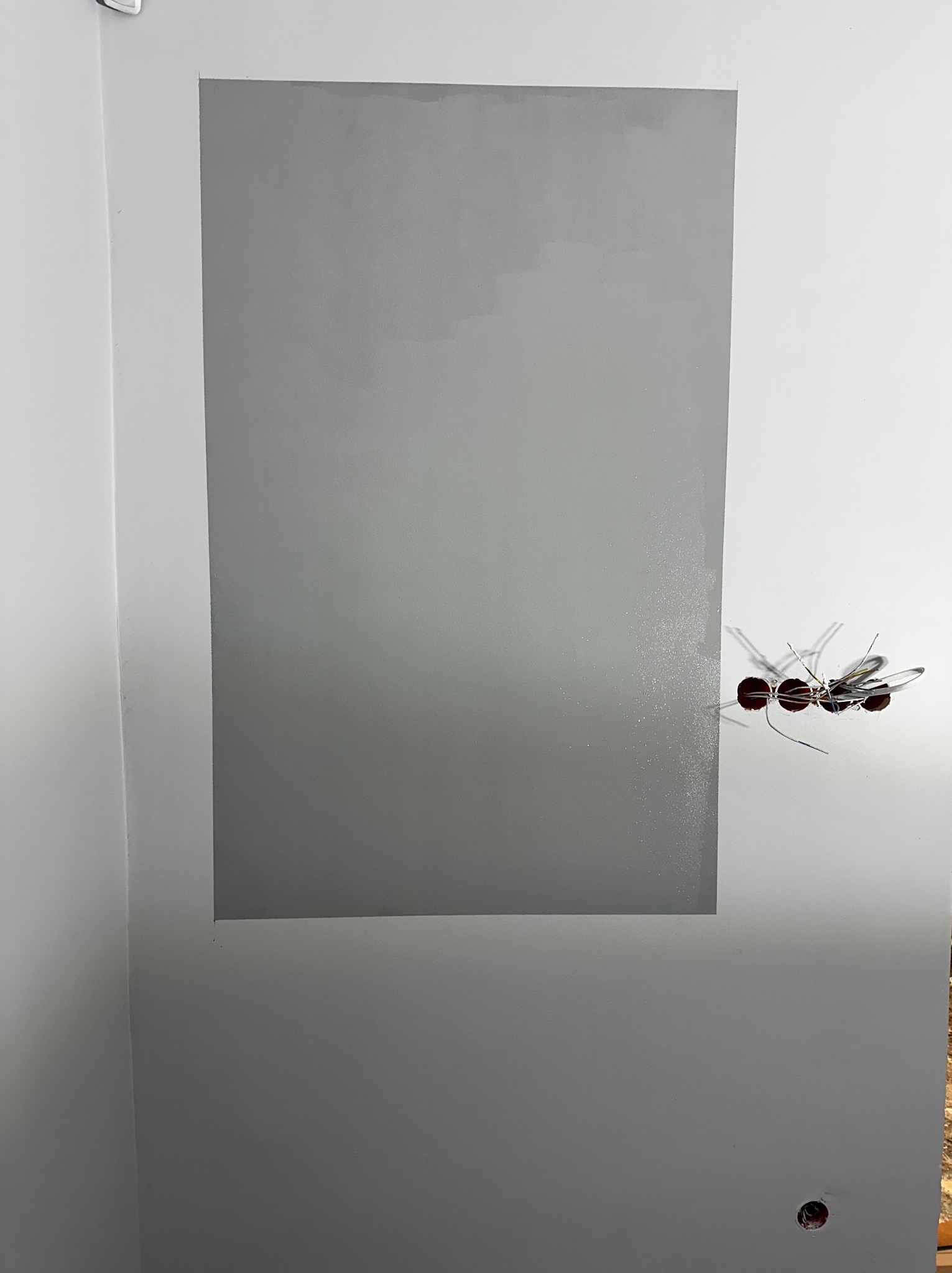 Świeżo pomalowany fragment ściany na szaro, obok wystające przewody elektryczne z puszki, widoczny otwór w dolnej części ściany.