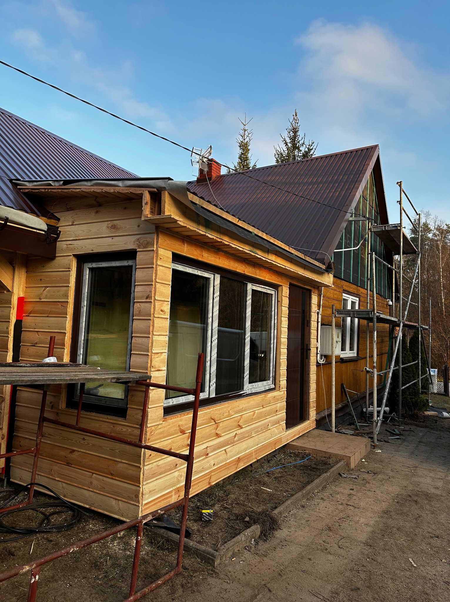 Drewniany dom w trakcie budowy z widocznymi rusztowaniami, brązowym dachem i nowymi oknami z białymi ramami, częściowo ocieplony, w tle drzewa i błękitne niebo.
