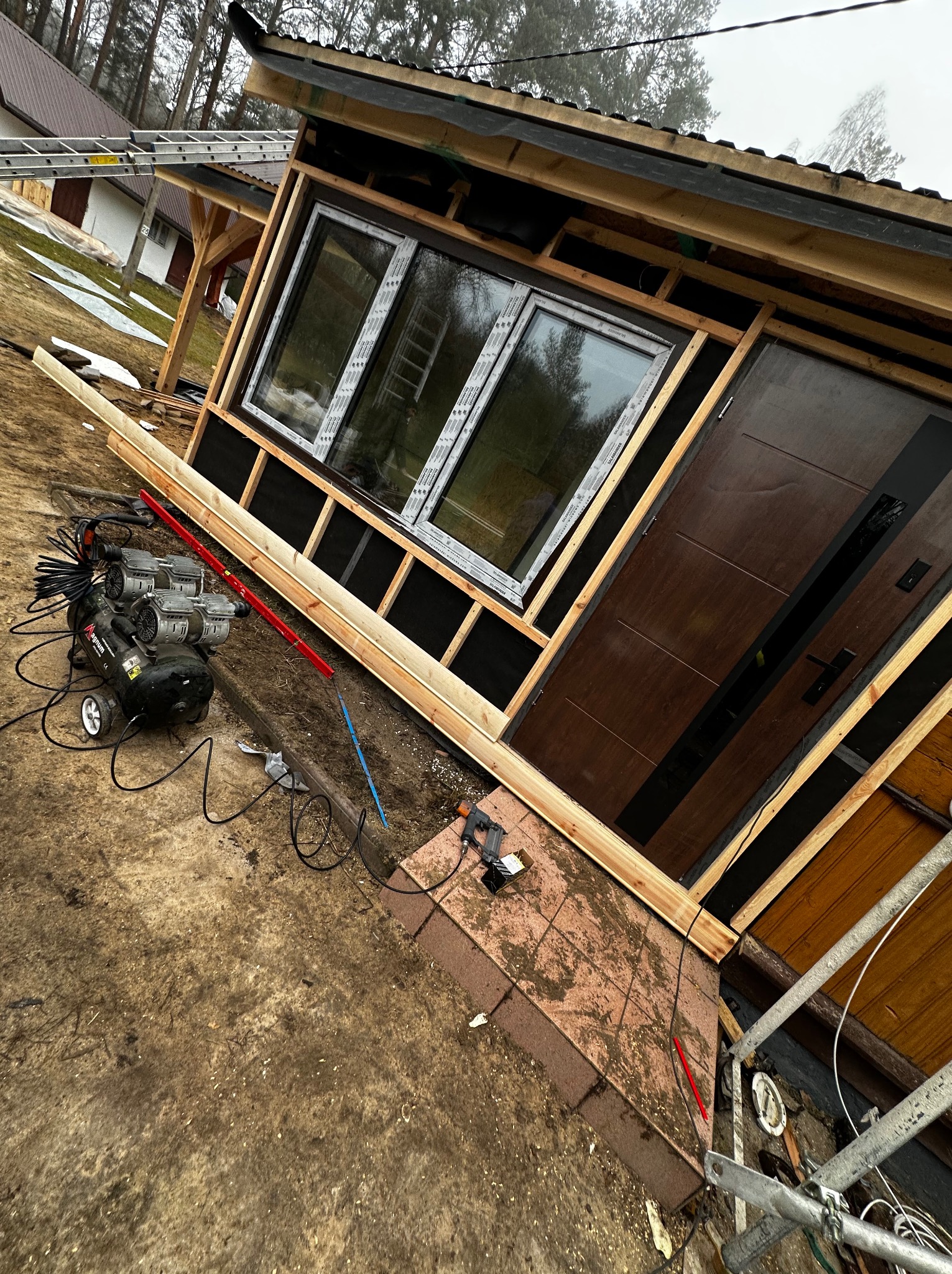 Montaż okna i drzwi wejściowych w drewnianej konstrukcji, widoczne narzędzia i kompresory na zewnątrz budynku.