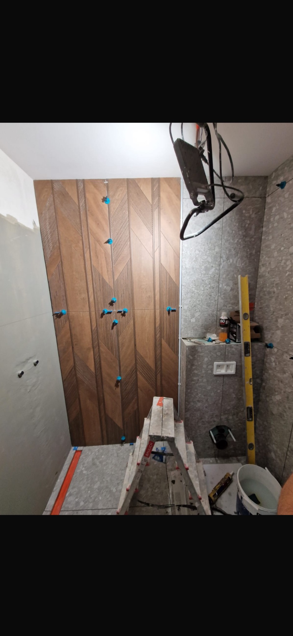 Montaż płytek drewnopodobnych w łazience, widoczne niebieskie krzyżyki dystansowe, poziomica i drabina.