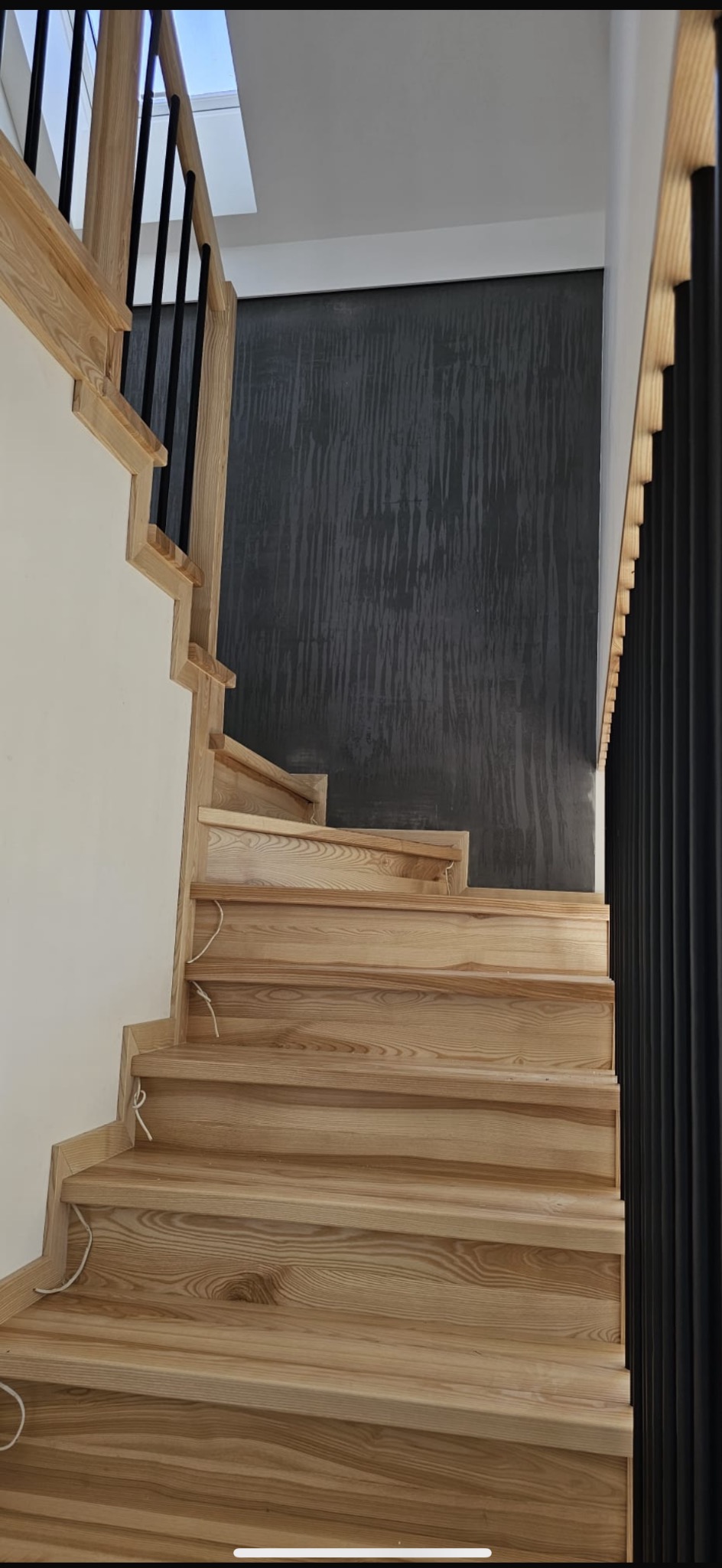 Jasne drewniane schody z czarną metalową balustradą na tle ściany z szarą dekoracyjną fakturą.