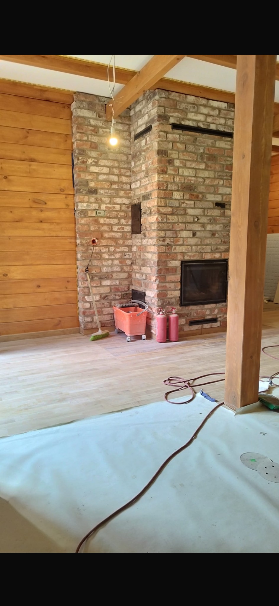Częściowo wykończone wnętrze z ceglanym kominkiem, drewnianymi panelami ściennymi i zabezpieczoną podłogą, widoczne narzędzia i akcesoria malarskie.