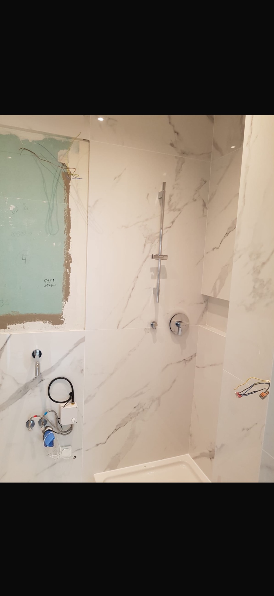 Łazienka w trakcie wykańczania: białe płytki imitujące marmur, zamontowana bateria prysznicowa, widoczne instalacje wodne i elektryczne przed zabudową, fragment brodzika, ściana przygotowana...