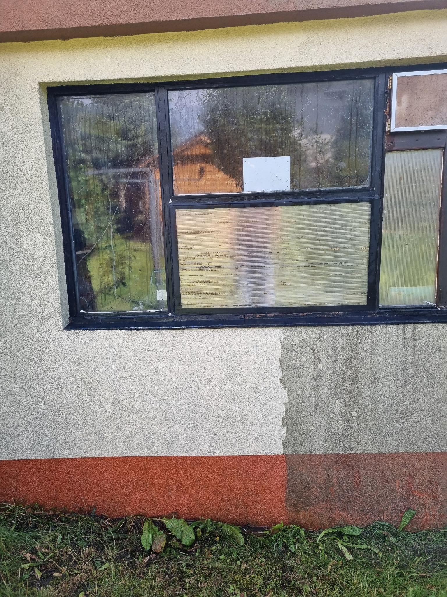 Stare okno z czarną ramą w elewacji budynku z widocznymi śladami wilgoci i zabrudzeń na ścianie. Częściowo odnowiona elewacja. Zarośnięta trawa u podstawy.