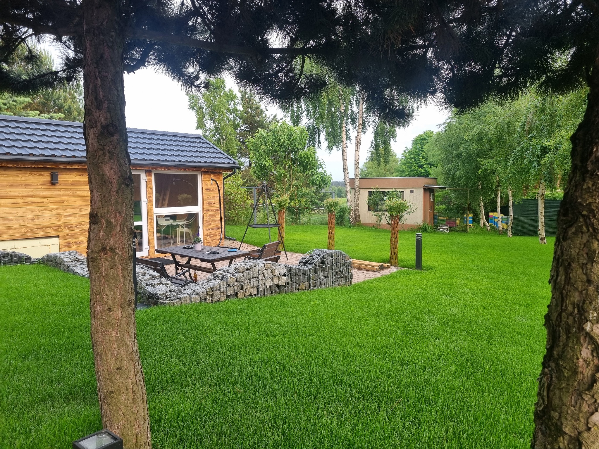 Zadbany ogród z równo przystrzyżonym trawnikiem, drewnianym domkiem i elementami architektury ogrodowej, takimi jak gabiony i oświetlenie. Widoczne drzewa i krzewy.