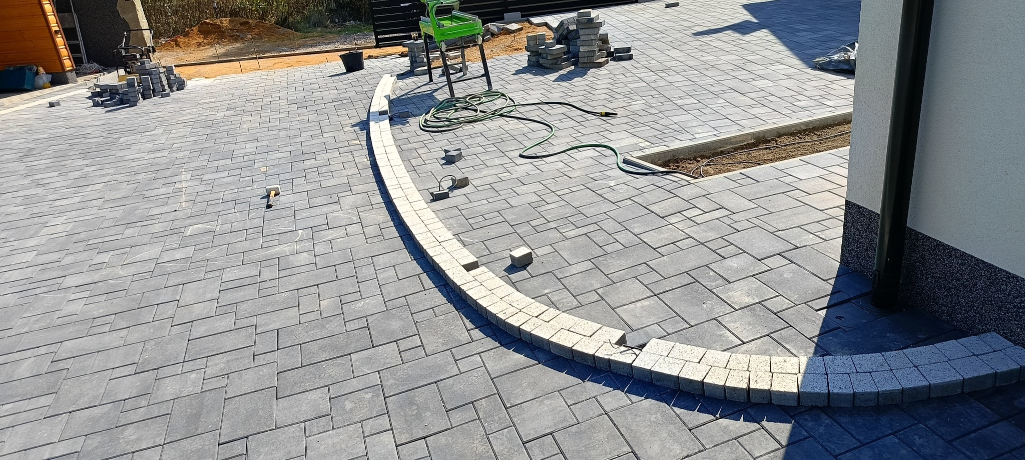 Szara kostka brukowa ułożona w nieregularny wzór, z jasną obwódką z kostki ułożonej łukiem, widoczny wąż ogrodowy, sterta kostki i narzędzia brukarskie.