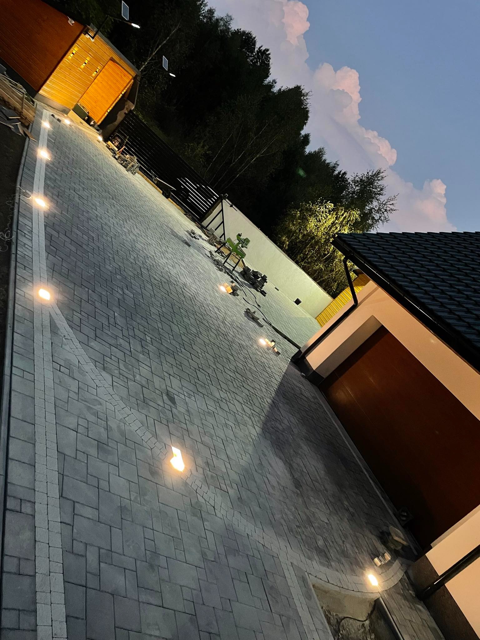 Oświetlona posesja z ułożoną szarą kostką brukową, widoczne lampy najazdowe, brama garażowa i fragment budynku w wieczornym oświetleniu.