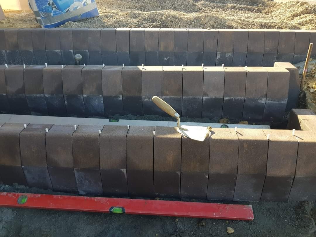 Montaż obrzeży z kostki brukowej w Chrzanowie, widoczna poziomica i kielnia, białe kołki stabilizujące, w tle worki z materiałem budowlanym.