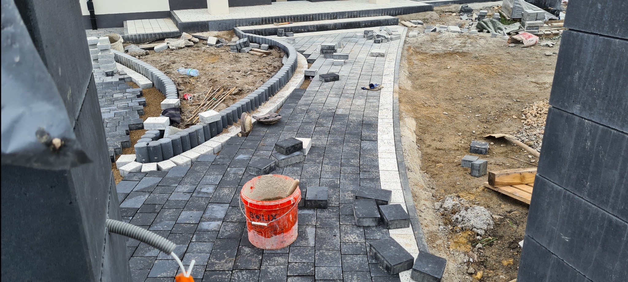 Budowa ścieżki z ciemnej kostki brukowej, widoczny czerwony kubeł z zaprawą, obrzeża z jasnej kostki, ziemia przygotowana pod dalsze prace. Ujęcie zza elementu betonowego ogrodzenia.
