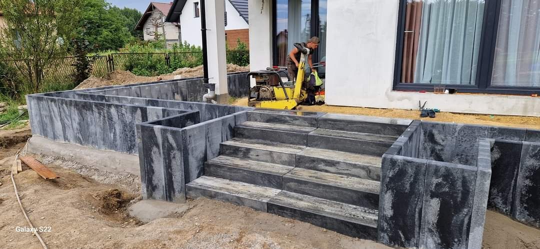 Budowa schodów wejściowych z kostki granitowej w odcieniach szarości, z widocznym zagęszczarką i pracownikiem w tle, na etapie przygotowania terenu.