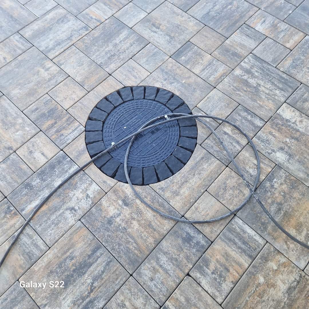 Ułożona kostka brukowa w odcieniach szarości i brązu z okrągłą studzienką kanalizacyjną z czarnym deklem i wystającym przewodem.