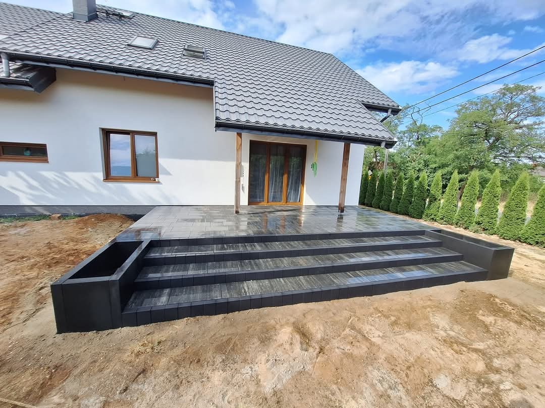 Nowoczesne schody z ciemnej kostki brukowej prowadzące do domu z białymi ścianami i szarą dachówką. Widoczne donice z kostki po bokach schodów.