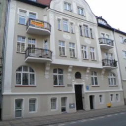 Odnowiona kamienica z beżową elewacją, białymi zdobieniami i balkonami z czarną balustradą. Na jednym z balkonów widoczna tabliczka 'Sprzedam'.