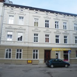 Mat-Bud Sp. z o.o. - Odnowiona fasada trzypiętrowej kamienicy z ozdobnymi elementami architektonicznymi, widok z poziomu ulicy, zaparkowany samochód i szyld salonu usługowego.