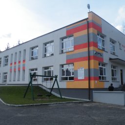 Mat-Bud Sp. z o.o. - Budynek przedszkola z pomalowaną elewacją w pasy szaro-pomarańczowo-czerwone, widoczne okna, huśtawka i schody wejściowe.