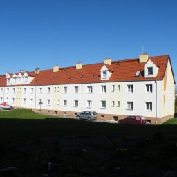 Mat-Bud Sp. z o.o. - Długi, trzypiętrowy budynek mieszkalny z czerwonym dachem, widoczny z perspektywy trawnika, z zaparkowanymi samochodami i łodzią na przyczepie w tle.