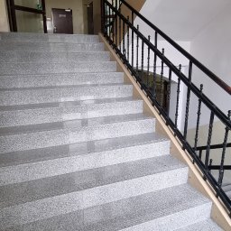 Mat-Bud Sp. z o.o. - Granitowe schody z czarną, kutą balustradą w budynku z beżowymi ścianami i łukowym przejściem w tle.